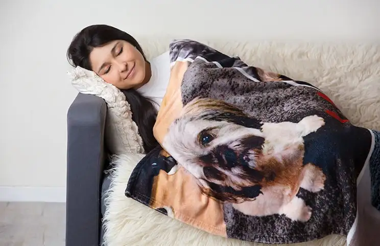 Una mujer envuelta en una manta de visón personalizada de Printerpix con diseño de perros. Ella está felizmente dormida en el sofá.