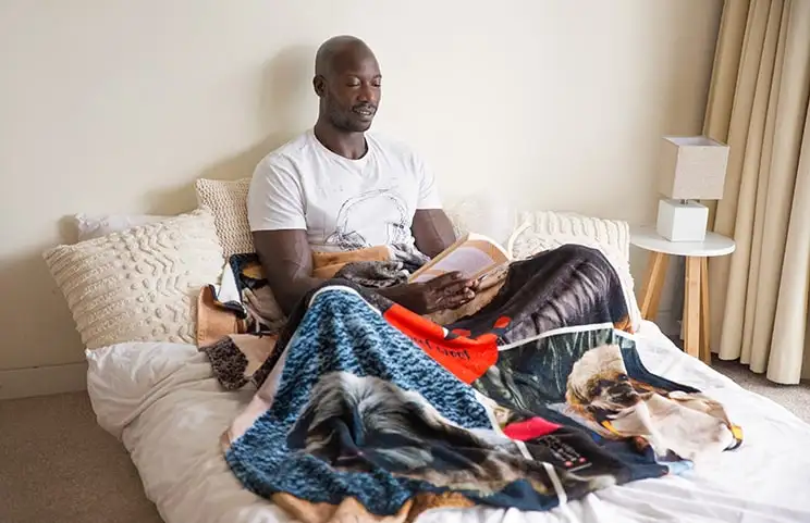 Un hombre lee con una manta personalizada de Printerpix con diseño de collage de perros sobre su regazo.